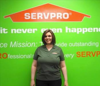 Female employee Heather Lyons standing in front of a green wall with the SERVPRO logo and mission statement below it.